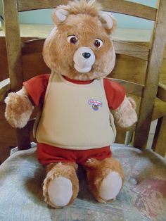 a brown teddy bear sitting on top of a chair wearing a bib and overalls