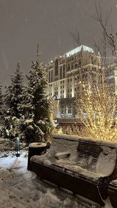 a park bench covered in snow next to trees and buildings at night with lights on