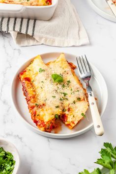 Two cheese-filled manicotti topped with melted mozzarella, fresh basil, and Parmesan, served on a white plate with a fork, alongside a baking dish and garnished parsley.