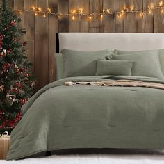 a bed covered in green sheets and pillows next to a christmas tree with lights on it