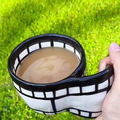 a hand holding a black and white bowl with brown liquid in it on top of green grass