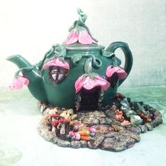 a green teapot with pink flowers on it sitting on top of rocks and pebbles