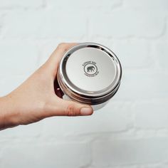 a hand holding a round metal container with an elephant on the lid and a white brick wall behind it