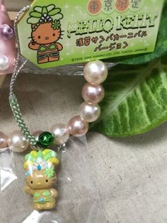 a hello kitty necklace with pearls and a green bow on it's head is sitting next to a leaf