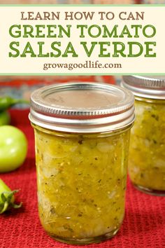 two jars filled with green tomato salsa sitting on top of a red cloth next to peppers