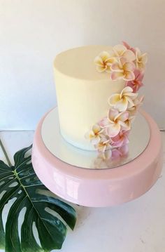 a white cake with pink and yellow flowers on top next to a green leafy plant