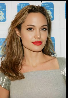 a woman with long brown hair and red lipstick wearing a grey top is posing for the camera