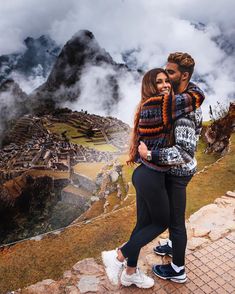 a man and woman standing on the edge of a mountain with clouds in the background