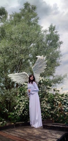 a woman with white wings standing on a wooden platform in front of trees and flowers