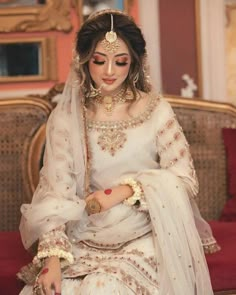 a woman in white and gold bridal outfit sitting on a couch with her hands clasped