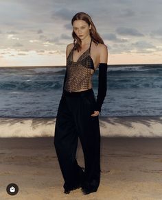 a woman standing on top of a sandy beach next to the ocean with her hands in her pockets