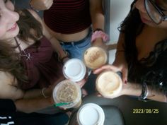 three girls are holding cups with drinks in them and looking at something on the table