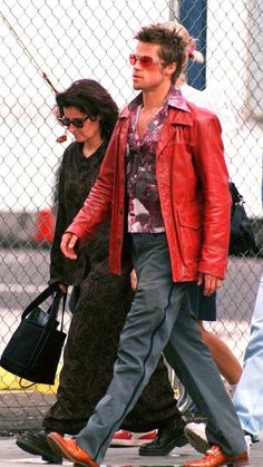 a man in a red jacket and sunglasses walking down the street with a woman behind him