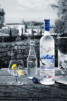 a bottle of vodka next to two glasses on a wooden table with an old stone wall in the background