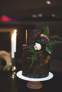 a three tiered cake with flowers and greenery sits on a table next to two candles