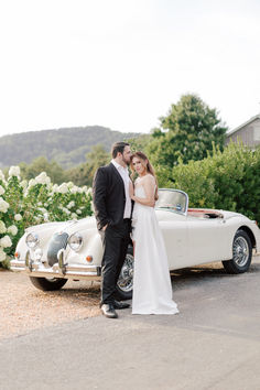 a bride and groom standing in front of a vintage car with white flowers behind them