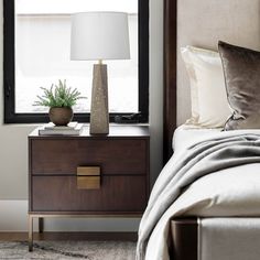 a bedroom with a bed, night stand and lamp next to the window that has a potted plant on it