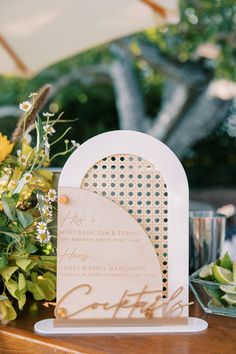 a wooden sign sitting on top of a table next to flowers and greenery with an umbrella in the background