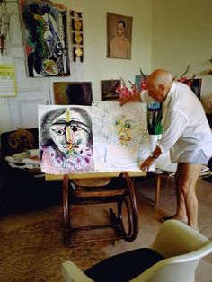 an older man is painting in his living room with paintings on the wall behind him