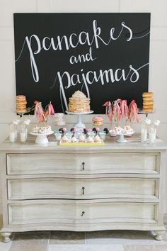 a dessert table with cakes and pastries on it in front of a chalkboard sign