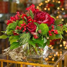 a vase filled with red flowers and greenery on top of a glass table next to a christmas tree