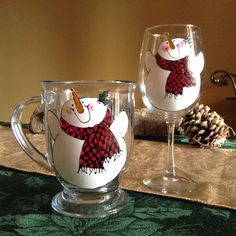 two wine glasses with snowmen painted on them sitting on a table next to pine cones