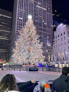 people looking at a large christmas tree in the middle of a city with tall buildings