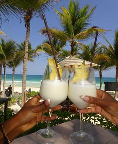 two people holding up glasses with drinks in front of palm trees and the beach behind them