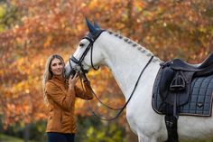 a woman standing next to a white horse in front of trees with orange leaves on it