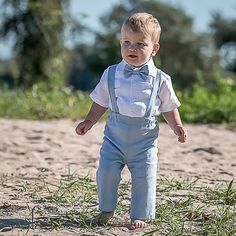 a little boy that is standing in the sand wearing a bow tie and suspenders