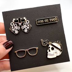 "A perfect pin pack for the optometrist or eye lover.  The pack includes four pins, 1) \"one or two?\", 2) silver, black, or gold spectacle frames, 3) skull with binocular indirect ophthalmoscope, and 4) phoropter. Choose the spectacles pin color when adding to your cart.  The set is priced as 4 pins for the price of 3! A great set of pins for your clinic coat, scrubs, or bag. Each pin is about 1\"." Optometry Aesthetic, Optician Training, Optician Marketing, Optometrist Office, Eyewear Store Design, Nurse Aesthetic, Bag Pins, Spectacles Frames, Eye Pins