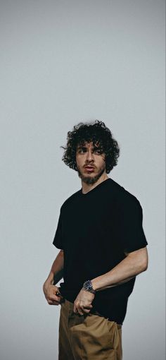 a man with curly hair standing in front of a white wall wearing a black t - shirt and khaki pants