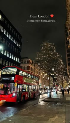 a red double decker bus driving down a street next to tall buildings with lights on