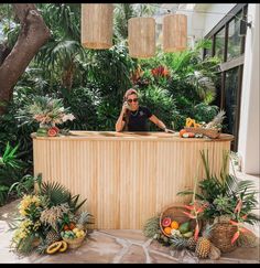 a woman standing behind a wooden counter surrounded by tropical plants and fruit in front of her