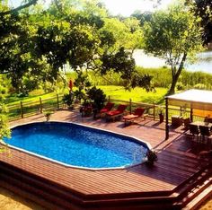 an above ground pool surrounded by decking and chairs next to a lake with trees in the background