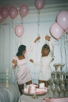 two women in white dresses holding pink balloons and standing next to cake on a table