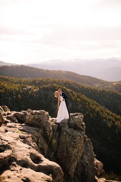 a bride and groom standing on top of a rocky cliff with mountains in the background