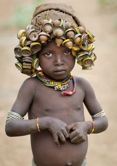 a young boy with beads on his head and bracelets around his neck, standing in the dirt