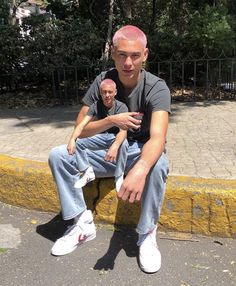 a man with pink hair sitting on a curb next to another man wearing white shoes