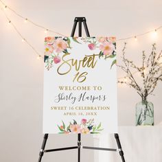 a welcome sign with flowers on it in front of a white wall and string lights