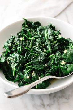 a white bowl filled with spinach on top of a marble counter next to a spoon