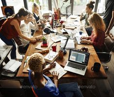 a group of people sitting around a wooden table working on laptops and writing notes