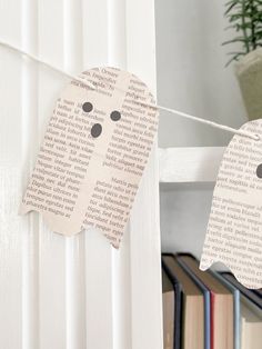 two paper ghost decorations hanging from a clothes line next to bookshelves and a potted plant