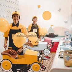 two men in yellow aprons stand behind a toy car with balloons on the back