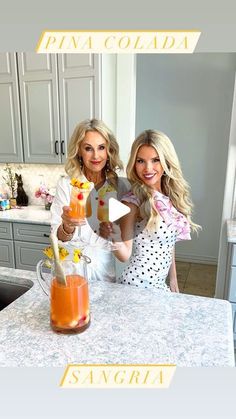 two women standing in front of a kitchen counter with drinks on the counter and one holding a blender