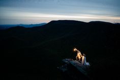 a bride and groom standing on top of a mountain at night with their arms in the air