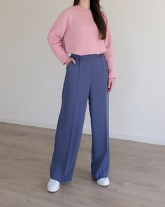 a woman standing in front of a white wall wearing blue pants and a pink sweater