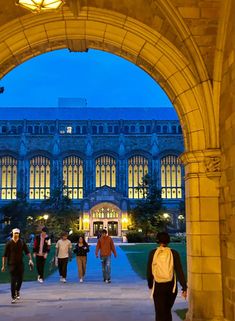 people walking under an archway in front of a large building at night with lights on