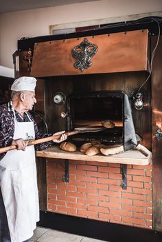 an old man holding a stick in front of a brick oven filled with loaves of bread