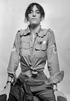 a woman sitting on the ground with her hands in her pockets, wearing an army uniform